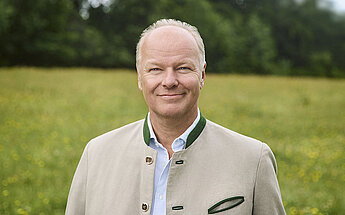 Ein Mann in einer beigen Jacke mit grünem Kragen steht in einem Feld mit gelben Blumen.