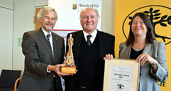Drei Personen, zwei Männer und eine Frau, halten eine Auszeichnung und eine Urkunde. Die Auszeichnung trägt den Namen "Okologija-Preis 2017", und die Urkunde ist aus Rheinland-Pfalz.
