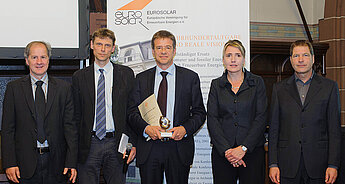 Eine Gruppe von fünf Personen, vermutlich bei einer Preisverleihung, von denen eine eine Trophäe in der Hand hält und im Hintergrund ein Banner mit dem Logo von "EUROSOLAR".