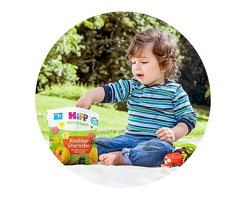 Ein Kleinkind, das im Freien sitzt und nach einer Snackpackung mit der Aufschrift "Hipp Bio Fruit Kinder" greift.