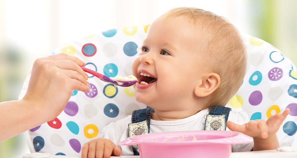 Ein fröhliches Baby, das von einem Erwachsenen mit einem Löffel gefüttert wird, sitzt in einem Hochstuhl mit einem bunten Tupfenmuster.