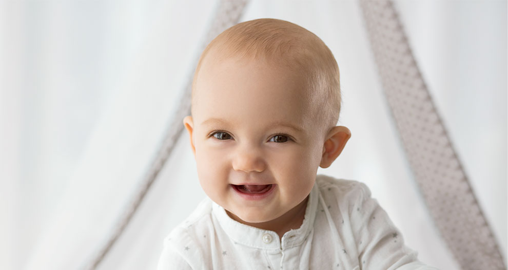 Ein Baby mit freudigem Gesichtsausdruck, das ein weißes Outfit trägt, vor dem Hintergrund eines weißen Vorhangs mit einem Muster.