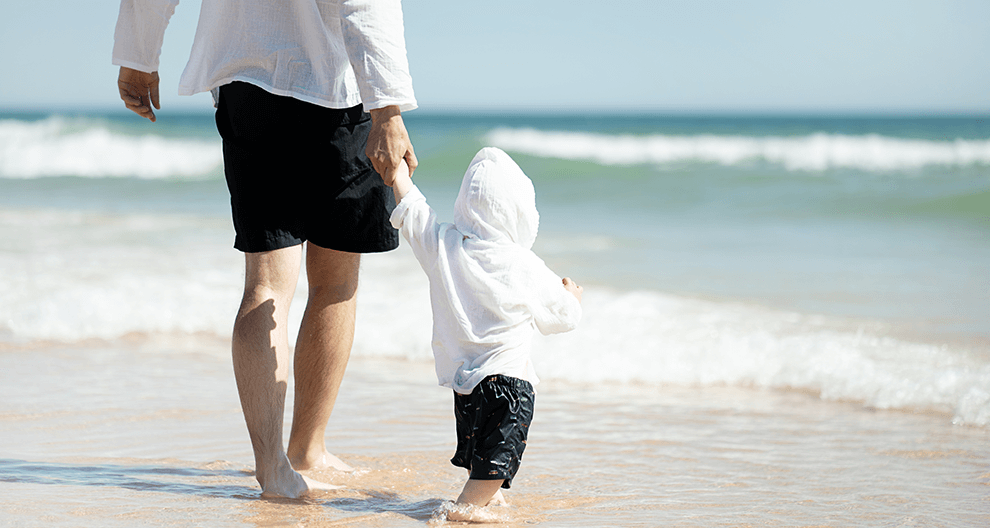 Ein Erwachsener und ein Kind spazieren Hand in Hand an einem Strand entlang, während im Hintergrund die Wellen sanft schlagen.