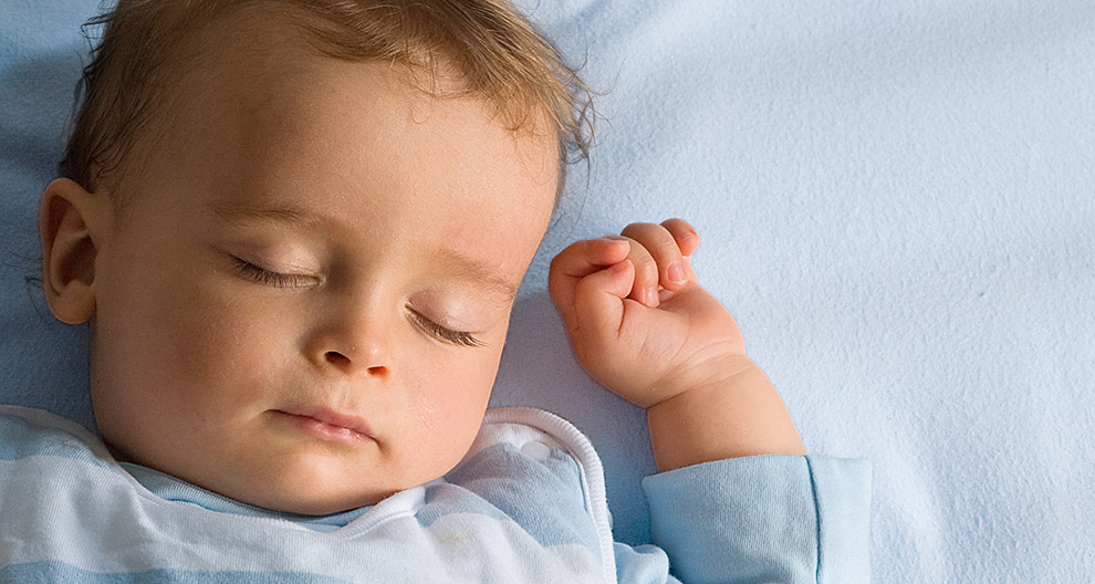 Ein scheinbar tief schlafendes Baby mit geschlossenen Augen und entspanntem Gesichtsausdruck, das auf einer blauen Fläche ruht.