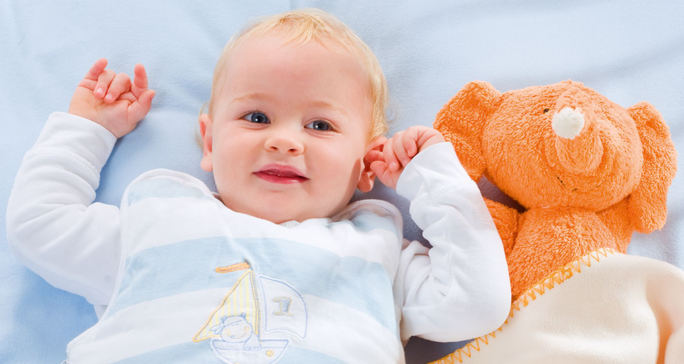 Ein Baby, das auf einer blauen Fläche liegt und ein weißes Outfit mit einem Segelbootmuster trägt, neben einem orangefarbenen Plüschtier, das einem Elefanten ähnelt.