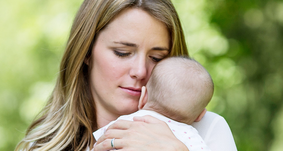 Eine Frau mit blondem Haar, die ein Baby an ihr Gesicht drückt, beide scheinen in einem ruhigen Moment zu sein.