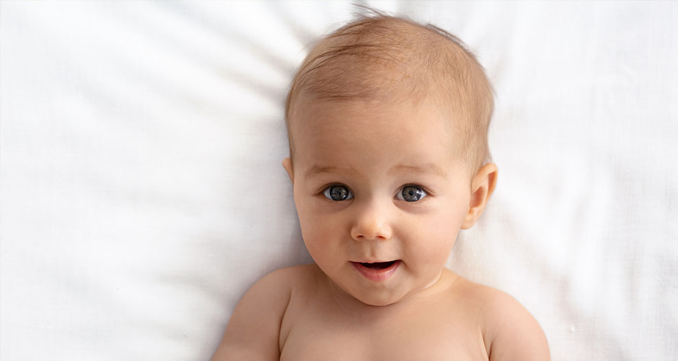 Ein Baby mit blauen Augen, das auf einer weißen Fläche liegt und direkt in die Kamera schaut.