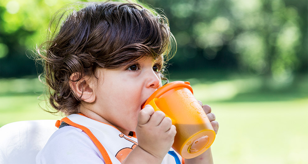Ein kleines Kind, vermutlich ein Kleinkind, nippt im Freien an einer orangefarbenen Schnabeltasse.