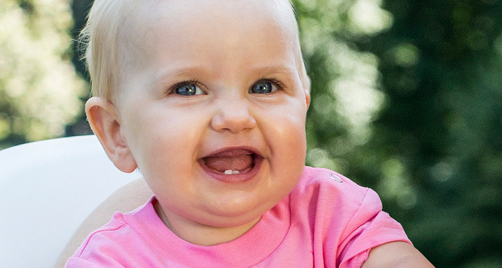 Ein Baby mit blondem Haar, das ein rosa Hemdchen trägt, lacht mit einem strahlenden Lächeln vor einem grünen Hintergrund.