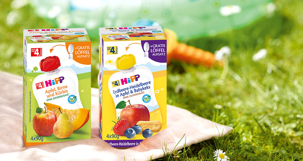 Zwei Fruchtsaftschachteln mit der Aufschrift "4Hipp" und verschiedenen Fruchtkombinationen, die auf einer Decke im Freien stehen, mit einer Karotte und Blumen im Hintergrund.