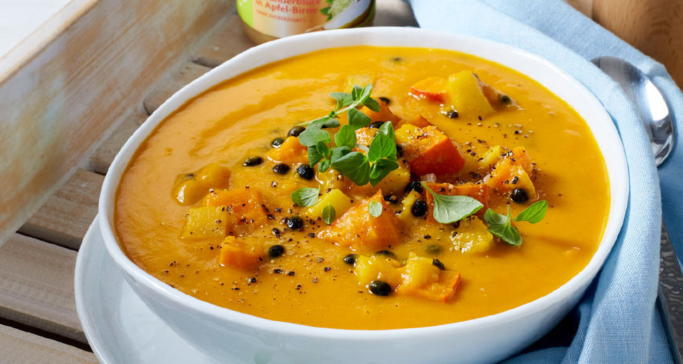 Eine Schüssel mit einer leuchtend orangefarbenen Suppe, garniert mit frischen Kräutern und Gewürzen, begleitet von einer Flasche Apfelsauce und einem Löffel.