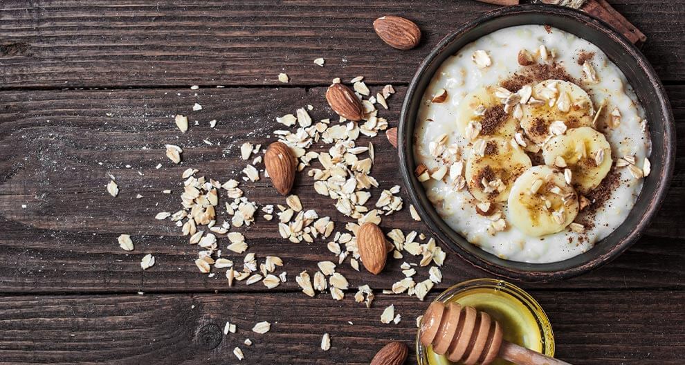 Eine Schüssel Haferflocken mit geschnittenen Bananen, Mandeln und Zimt, begleitet von einem Honiglöffel und gestreuten Haferflocken auf einem rustikalen Holzuntergrund.