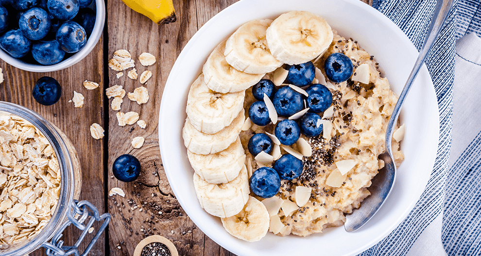 Eine Schüssel Haferflocken mit geschnittenen Bananen, Blaubeeren und Chiasamen, dazu ein Glas Haferflocken und eine Banane.