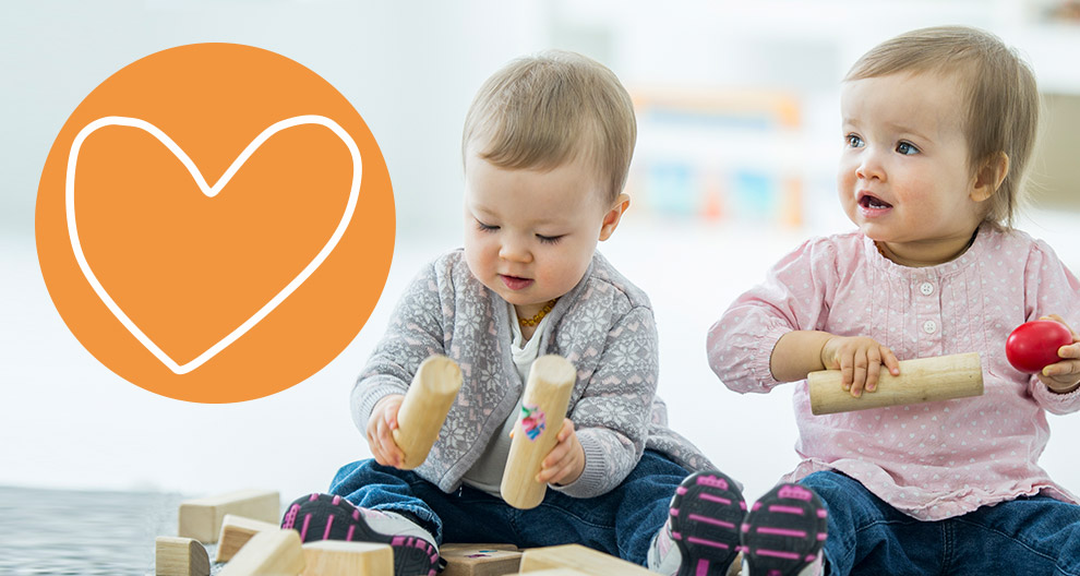 Zwei Kleinkinder spielen mit Holzklötzen, von denen eines ein rotes Objekt in der Hand hält, vor einem orangefarbenen Hintergrund mit einem weißen Herzsymbol.