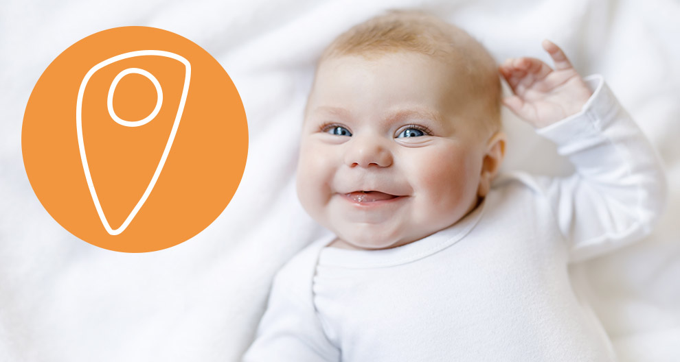 Ein Baby mit blauen Augen, das ein weißes Outfit trägt und auf einer weißen Fläche neben einem orangefarbenen Kreis mit einem weißen Stecknadelsymbol liegt.